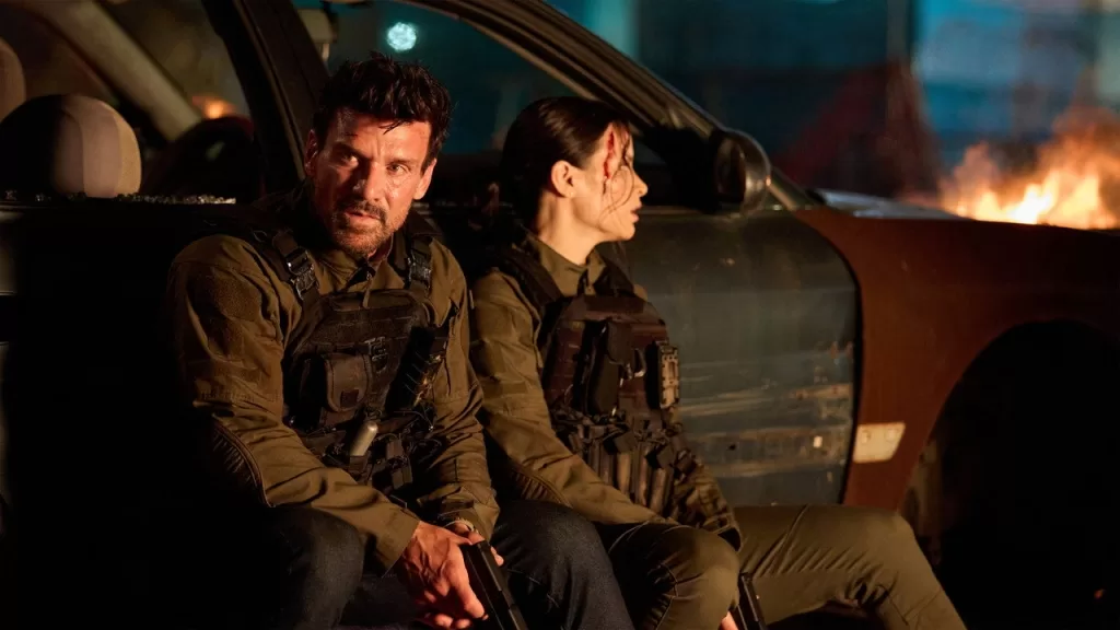 Frank Grillo e outro personagem armados se protegem atrás de um carro à noite, com fogo ao fundo, em cena de A Noite dos Lobisomens