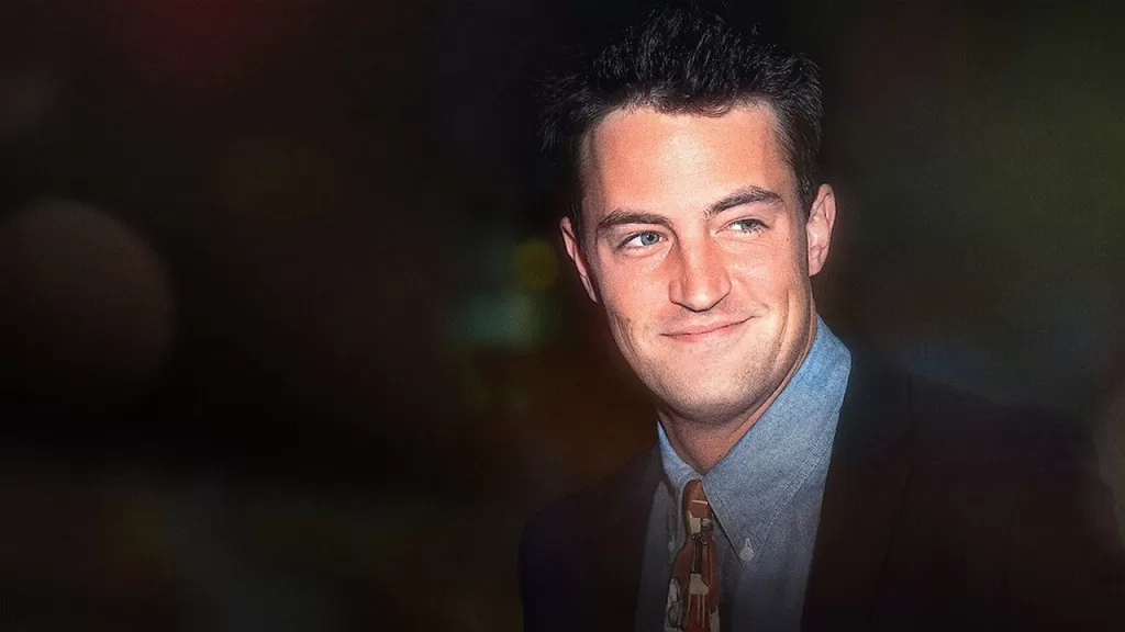 Matthew Perry sorri de terno escuro e camisa azul em imagem de divulgação da série documental Aquele Sobre Matthew Perry