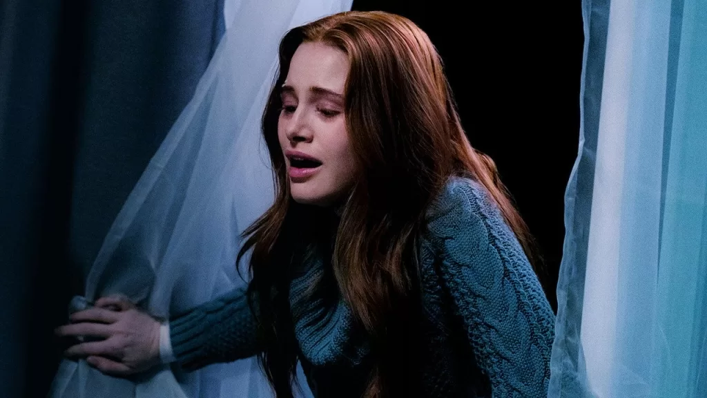 Madelaine Petsch como Ellen, assustada, encostada em uma janela com cortinas em um apartamento