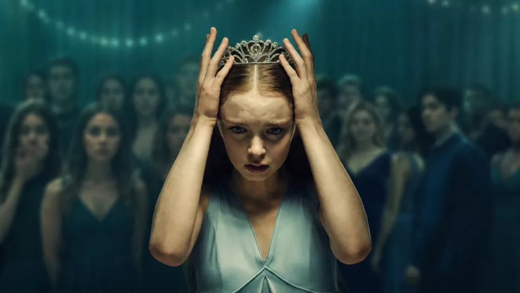 Summer H. Howell como Carrie White, com uma coroa na cabeça e as mãos no rosto, cercada por alunos no baile da escola