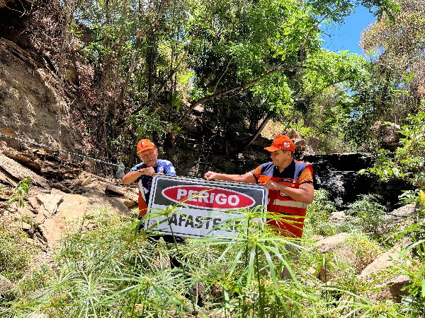 Dois agentes da Defesa Civil do Crato, de colete laranja, seguram placa de "Perigo – Afaste-se" na parte superior da Cascata, entre pedras e vegetação