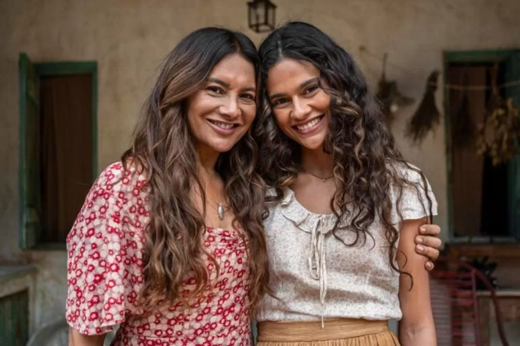 As atrizes Dira Paes e Arianne Botelho abraçadas e sorrindo em cenário de casa simples do sertão