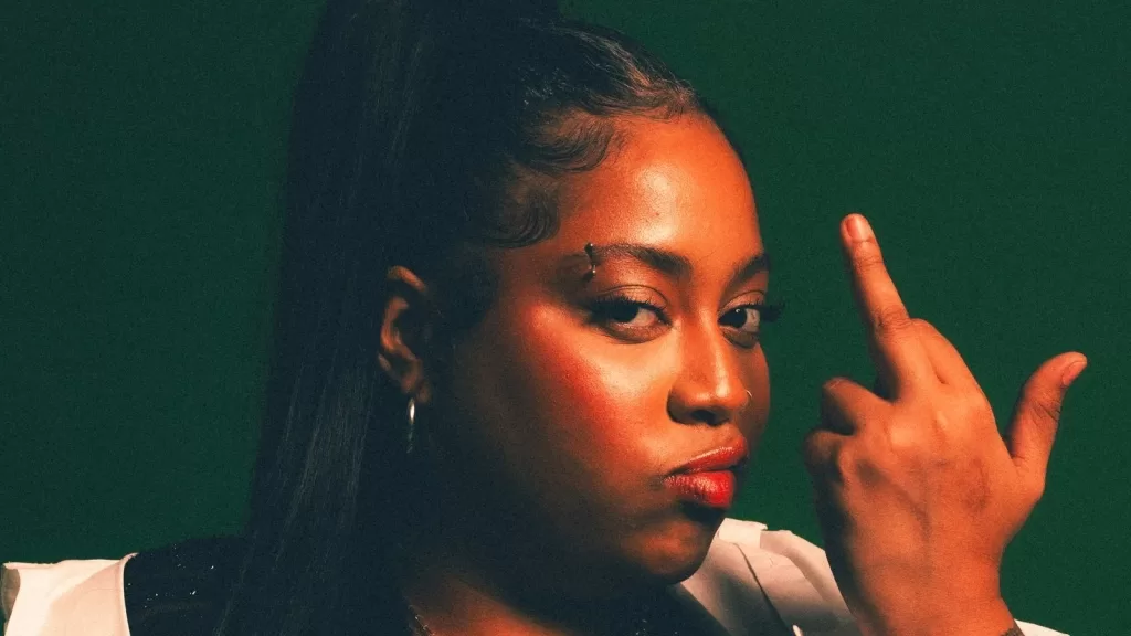 Retrato da DJ Bonekinha Iraquiana, de cabelo preso, com a mão próxima ao rosto, diante de fundo verde-escuro