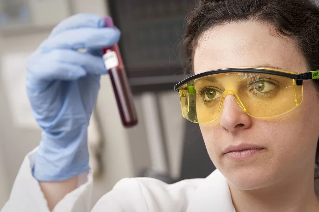 Pesquisadora de óculos de proteção e luvas azuis observa um tubo com amostra de sangue em laboratório