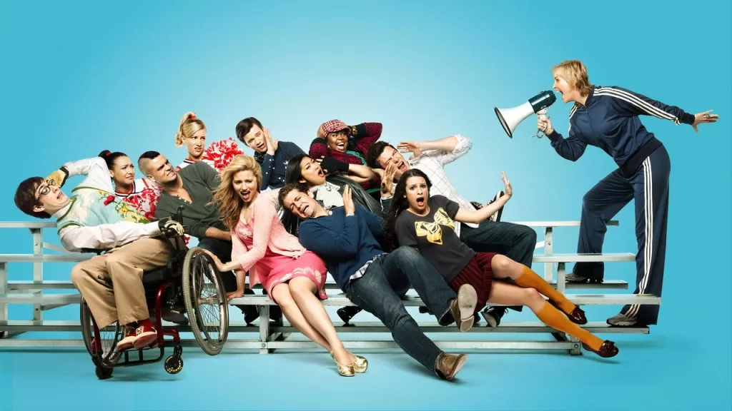 Elenco da série Glee posa em uma arquibancada com fundo azul, com o professor Will Schuester e a treinadora Sue Sylvester à direita