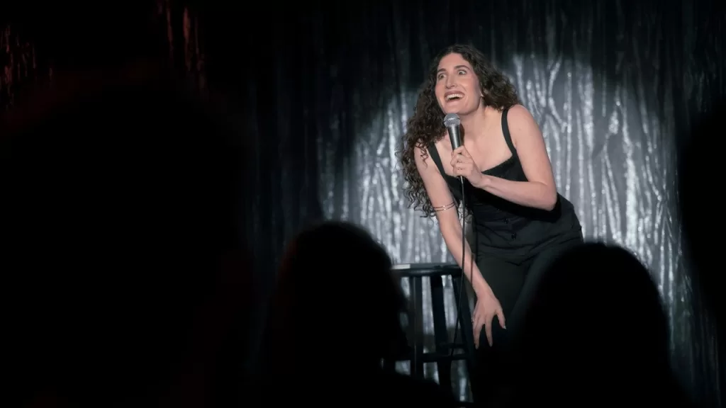 Kate Berlant se apresenta ao microfone em um palco com cortina prateada, diante da plateia