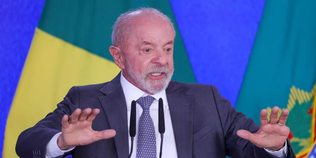 O presidente Luiz Inácio Lula da Silva discursa com as mãos abertas diante de microfones, com a bandeira do Brasil ao fundo