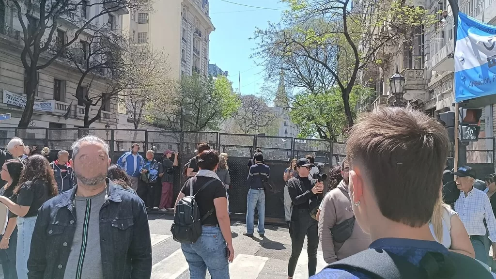 Manifestantes caminham por rua arborizada de Buenos Aires durante marcha em defesa das universidades públicas, com bandeira da Argentina à direita
