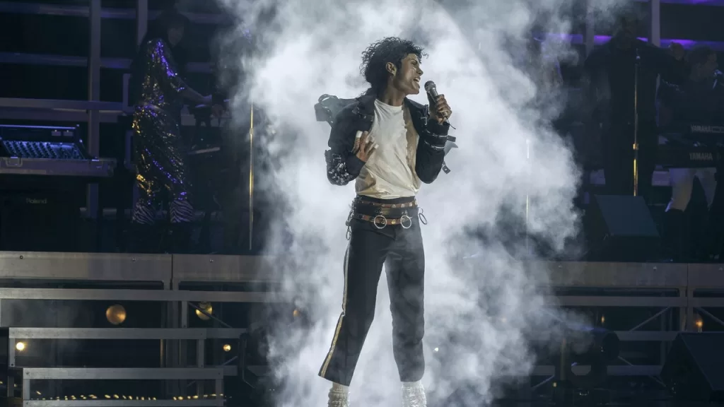 Jaafar Jackson como Michael Jackson, de jaqueta preta e camisa branca, cantando ao microfone em um palco tomado por fumaça e luzes