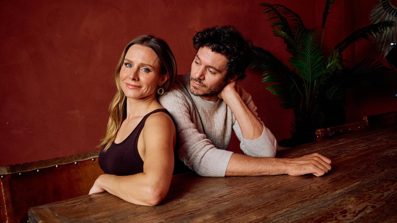 Kristen Bell e Adam Brody, protagonistas da série Ninguém Quer, sentados lado a lado em um ambiente de paredes vermelhas