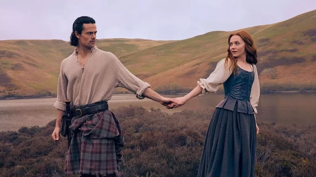 Casal de mãos dadas caminha por um campo nas Terras Altas da Escócia, com colinas ao fundo, em cena de Outlander: Blood of My Blood