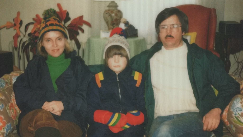 Foto de família antiga da comediante Maria Bamford ainda criança, ao lado dos pais e da irmã em uma sala decorada