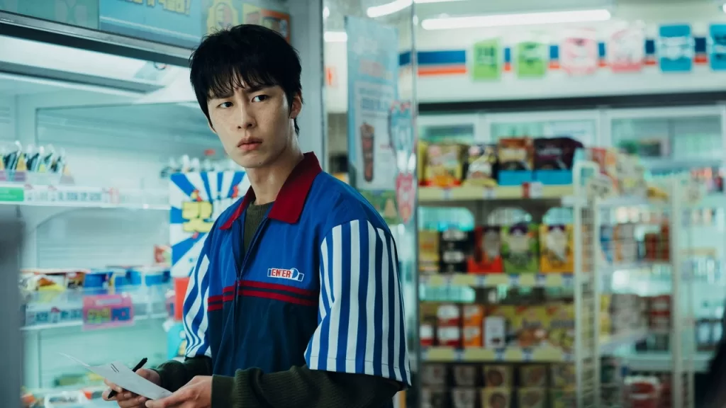 Lee Jae-wook com uniforme de funcionário entre as prateleiras de uma loja de conveniência, em cena da série Um Trabalho do Inferno